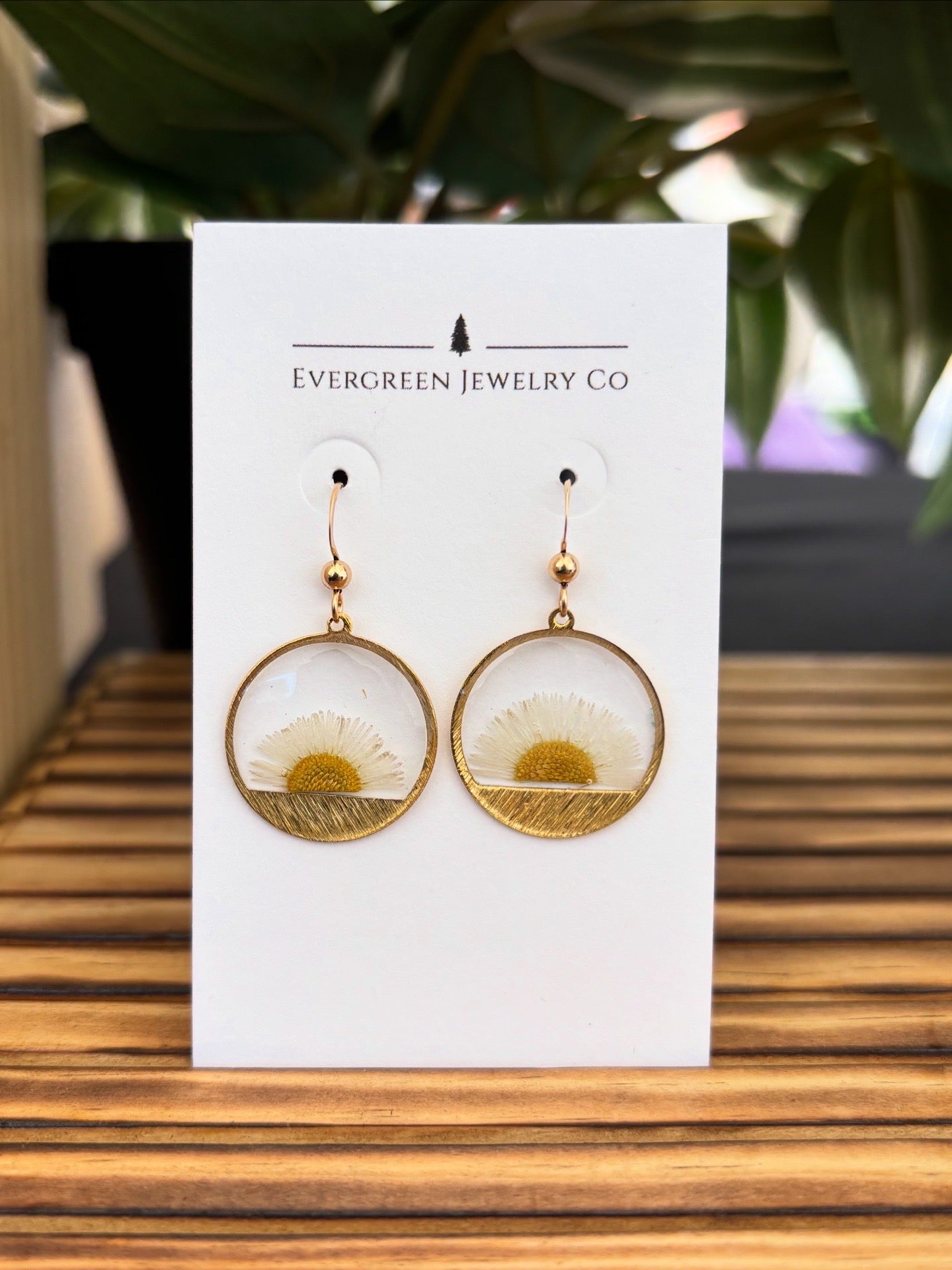 Gold earrings with floral design on a white card with 'Evergreen Jewelry Co' branding, placed on a wooden surface.