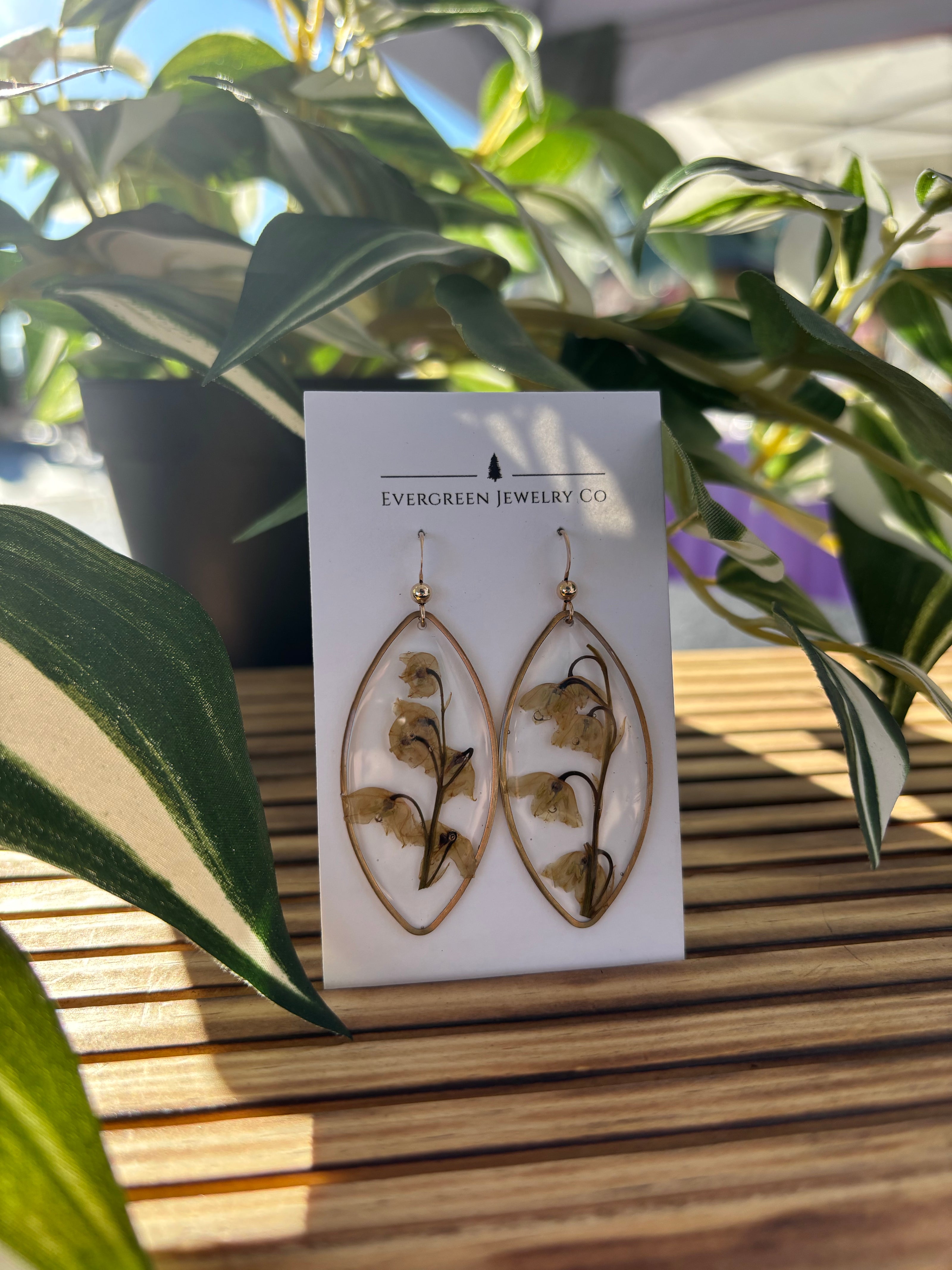 Pair of earrings made of clear resin and dried lily of the valley flowers on a card with green leaves in the background
